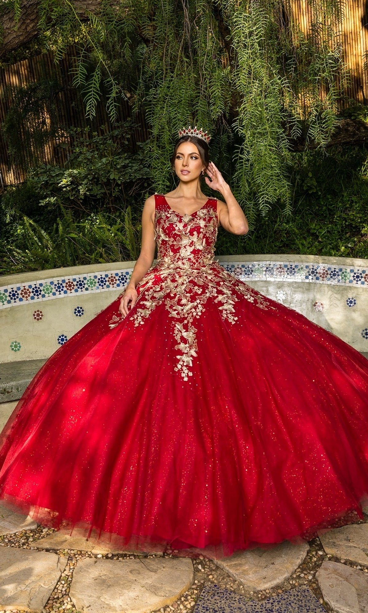 A young woman wearing the Quinceanera Ball Gown 8046J with metallic embroidery and a tiara stands outdoors by a decorative tiled bench amid lush greenery.